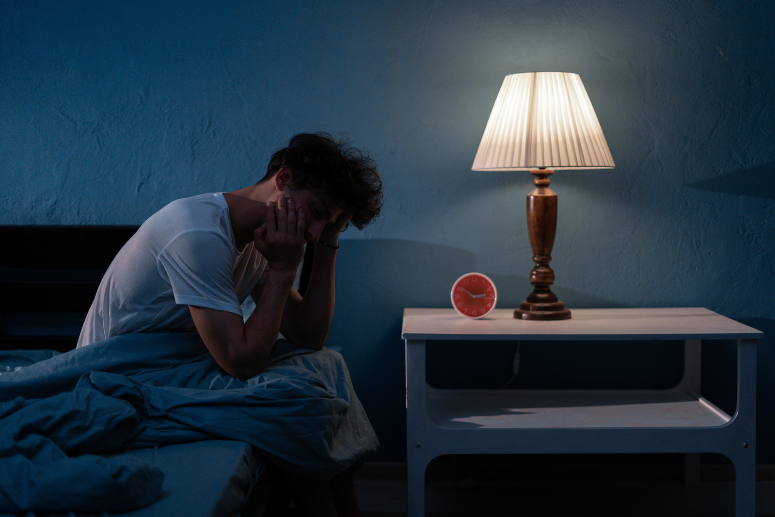 man holding head in pain while sitting on bed in the dark